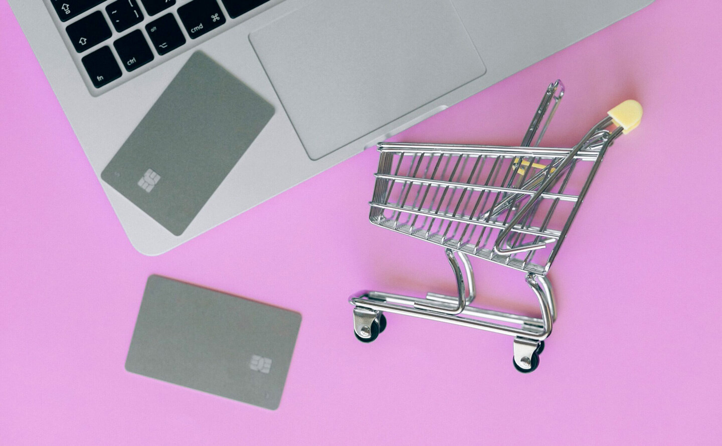 Computer su sfondo rosa con carrello della spesa appoggiato al tabolo che rappresenta lo shopping online